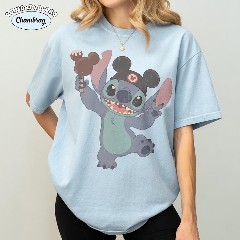 May include: A light blue short-sleeve t-shirt featuring a graphic of the character Stitch wearing black mouse ears and holding a brown chocolate treat. The text Comfort Colors and Chambray appears in the upper left corner of the image.