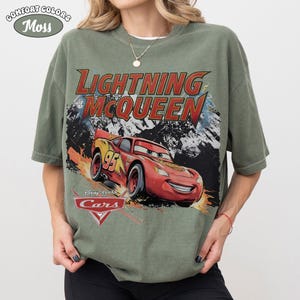 May include: A moss green short-sleeve t-shirt featuring a graphic print of the character Lightning McQueen from the movie Cars. The text Lightning McQueen is printed in red above the car, with the Disney Pixar Cars logo displayed below the car.