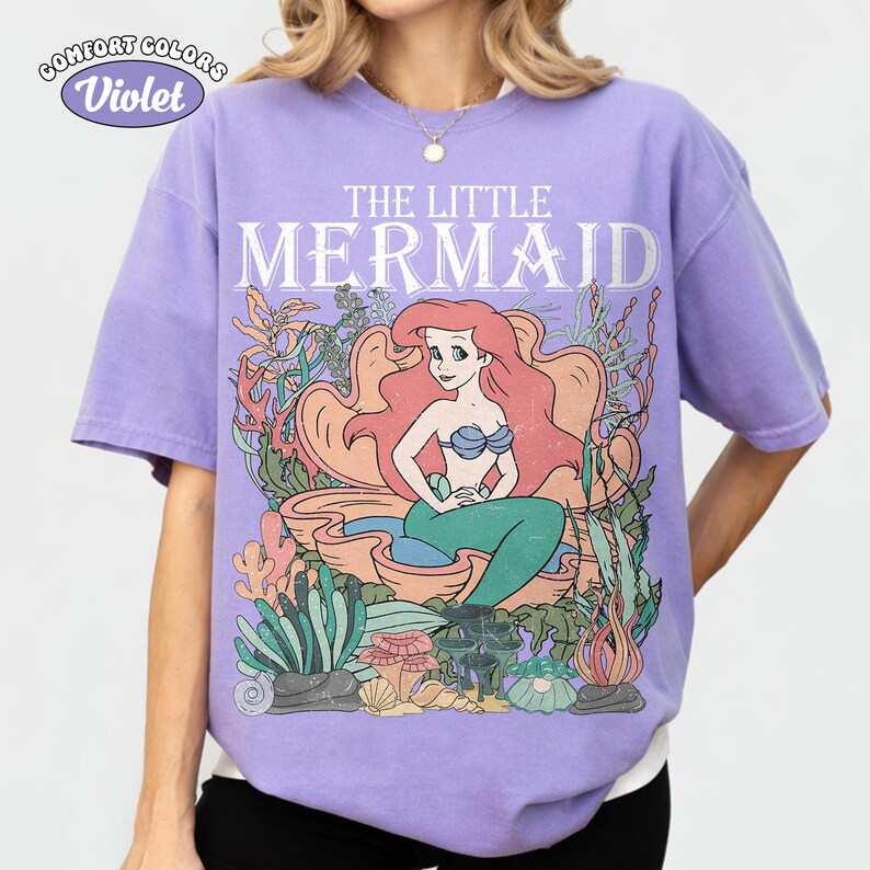 May include: Violet Comfort Colors t-shirt featuring a graphic of Ariel from The Little Mermaid. The design includes Ariel with red hair, a blue top, and a green tail, surrounded by coral and seaweed. The text "THE LITTLE MERMAID" is above the image.