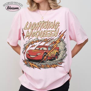 May include: Light pink t-shirt featuring a graphic of Lightning McQueen from the Cars movie. The design includes the text "LIGHTNING McQUEEN" and the number "95" on the car. The shirt has the "Comfort Colors" and "Blossom" logos.