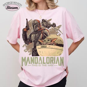 May include: A light pink t-shirt featuring a graphic print of a Mandalorian character holding a blaster, a small green creature in a floating pod, and a spaceship. The text "MANDALORIAN THIS IS THE WAY" is printed in green at the bottom of the graphic.