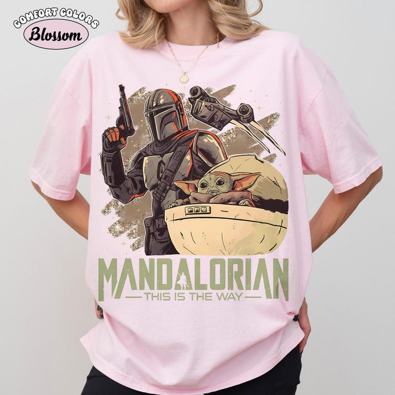 May include: A light pink t-shirt featuring a graphic print of a Mandalorian character holding a blaster, a small green creature in a floating pod, and a spaceship. The text "MANDALORIAN THIS IS THE WAY" is printed in green at the bottom of the graphic.