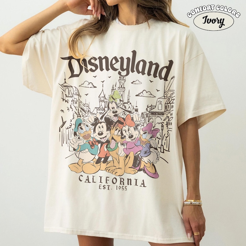May include: Ivory-colored t-shirt with a Disneyland graphic. The design features Mickey Mouse, Donald Duck, Goofy, Minnie Mouse, and Daisy Duck in front of a Disneyland park illustration. The text "Disneyland California Est. 1955" is also printed on the shirt.