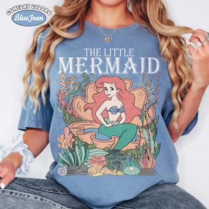 May include: Blue t-shirt with a graphic of Ariel from The Little Mermaid. The design features Ariel with red hair, a blue seashell bra, and a green tail, seated in a seashell. The text "THE LITTLE MERMAID" is above the image. The shirt has the Comfort Colors and Blue Jean logo.