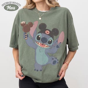 May include: A moss green short-sleeve t-shirt featuring a graphic of the character Stitch wearing black mouse ears and holding a brown chocolate-covered ice cream bar. The shirt has a crew neckline and a relaxed fit with a vintage-style print.