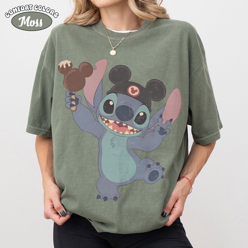 May include: A moss green short-sleeve t-shirt featuring a graphic of the character Stitch wearing black mouse ears and holding a brown chocolate-covered ice cream bar. The shirt has a crew neckline and a relaxed fit with a vintage-style print.