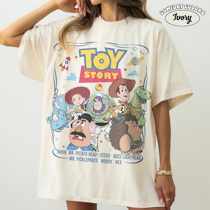 May include: Ivory-colored t-shirt featuring a colorful graphic of Toy Story characters. The design includes the words "TOY STORY" in a playful font, along with character names like Woody, Buzz Lightyear, and Jessie. The shirt is labeled "COMFORT COLORS" and "Ivory".