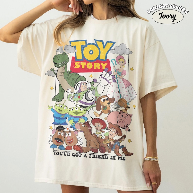 May include: An ivory t-shirt featuring a graphic print of Toy Story characters including Woody, Buzz Lightyear, Rex, and Slinky Dog. The design includes the text "TOY STORY" and "YOU'VE GOT A FRIEND IN ME" with a Comfort Colors Ivory label in the corner.
