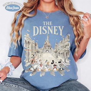 May include: Blue Comfort Colors t-shirt with "THE DISNEY" text and a graphic of Disney characters crossing a street in front of a castle. The shirt features Goofy, Daisy Duck, Donald Duck, Mickey Mouse, and Minnie Mouse.