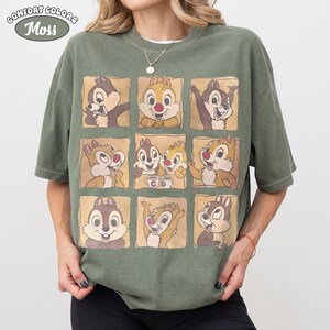 May include: Moss green Comfort Colors t-shirt with a grid of nine cartoon chipmunk images. The chipmunks are in different poses, set against a light brown background. The shirt has short sleeves and a relaxed fit. The Comfort Colors logo is in the upper left corner.