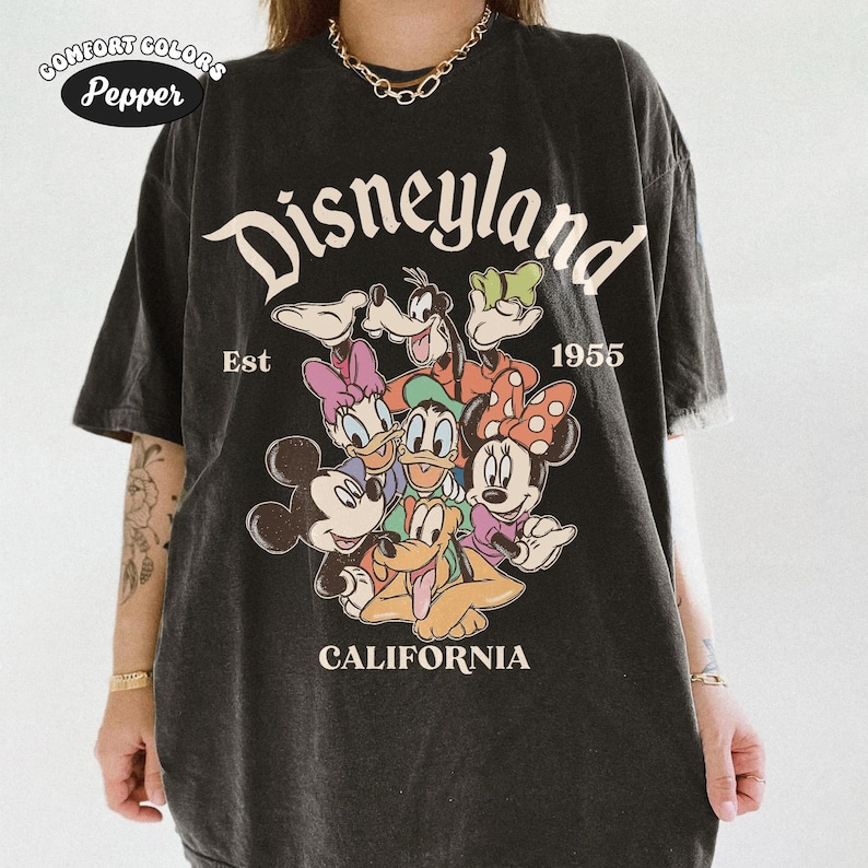 May include: Black t-shirt featuring a Disneyland graphic with Mickey Mouse, Minnie Mouse, Donald Duck, Daisy Duck, Goofy, and Pluto. The text "Disneyland California Est 1955" is printed on the shirt. The shirt is a Comfort Colors brand.