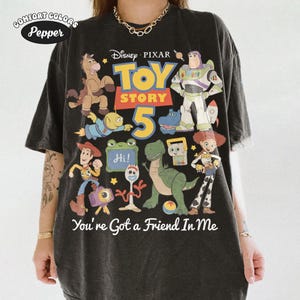 May include: Black Comfort Colors t-shirt featuring a colorful graphic of Toy Story characters, including Woody, Buzz Lightyear, and Rex. The text "TOY STORY 5" is prominently displayed, along with "You're Got a Friend In Me."