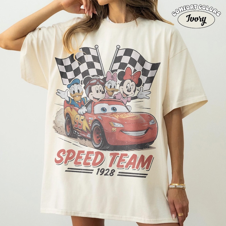 May include: Ivory t-shirt with a graphic of Lightning McQueen and Disney characters. The design includes checkered flags and the words "SPEED TEAM 1928". The shirt is a Comfort Colors brand.