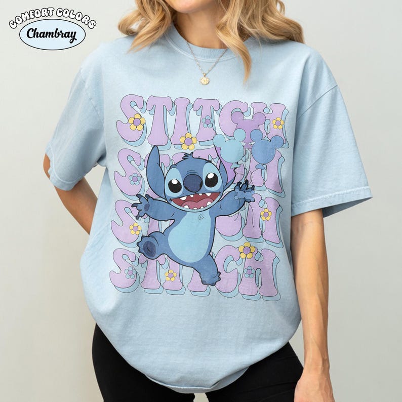 May include: A light blue short-sleeve t-shirt featuring a graphic of the character Stitch holding blue balloons. The word STITCH is repeated in large purple letters behind the character, accented with small yellow and purple flowers throughout.