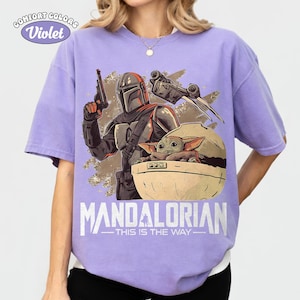 May include: A light purple short-sleeve t-shirt featuring a graphic print of a Mandalorian character holding a blaster and a small green creature in a floating pod. The text reads COMFORT COLORS Violet, MANDALORIAN, and THIS IS THE WAY.
