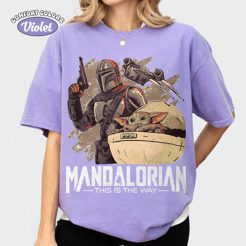 May include: A light purple short-sleeve t-shirt featuring a graphic print of a Mandalorian character holding a blaster and a small green creature in a floating pod. The text reads COMFORT COLORS Violet, MANDALORIAN, and THIS IS THE WAY.