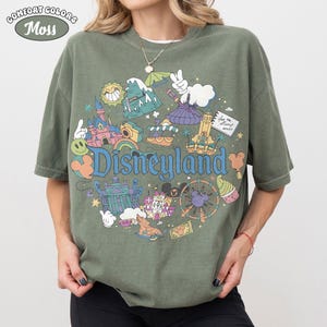 May include: A moss green t-shirt featuring a vibrant Disneyland graphic. The design includes a castle, a Ferris wheel, and the Disneyland logo in blue. The shirt has short sleeves and a loose fit, perfect for casual wear.