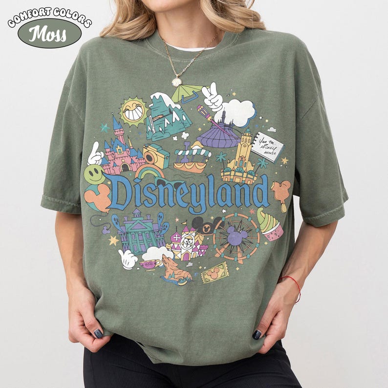 May include: A moss green t-shirt featuring a vibrant Disneyland graphic. The design includes a castle, a Ferris wheel, and the Disneyland logo in blue. The shirt has short sleeves and a loose fit, perfect for casual wear.