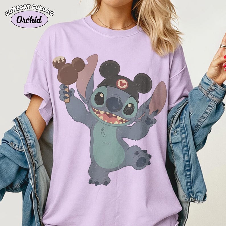 May include: A light purple short-sleeve t-shirt featuring a graphic of the character Stitch wearing black mouse ears and holding a chocolate-covered treat. The text Comfort Colors and Orchid is printed in the upper left corner of the garment.