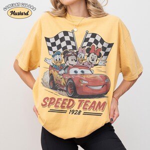 May include: Mustard-coloured t-shirt with a graphic of Disney characters in a red race car and chequered flags. The text "SPEED TEAM 1928" is printed below the car. The shirt has the words "Comfort Colors" and "Mustard" printed on the top left.