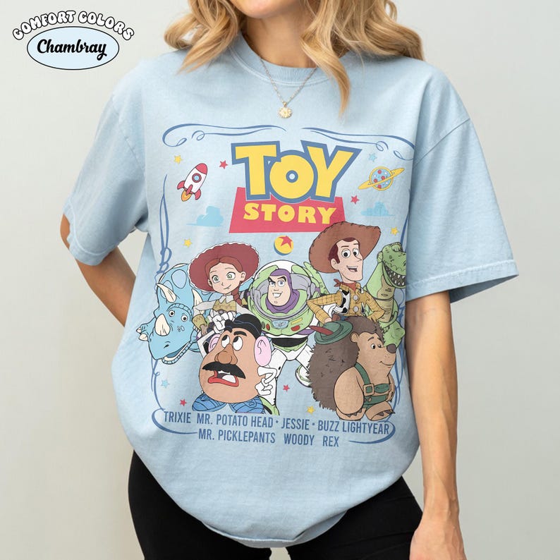 May include: Light blue Comfort Colors Chambray t-shirt featuring a graphic of Toy Story characters and the words "TOY STORY" in yellow. The shirt includes images of Woody, Buzz Lightyear, and other characters from the movie.