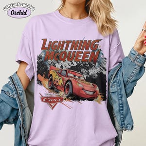 May include: A light purple short-sleeve t-shirt featuring a graphic of the red race car Lightning McQueen from the movie Cars. The text reads Lightning McQueen and includes the Cars logo. A Comfort Colors Orchid label is visible in the corner.