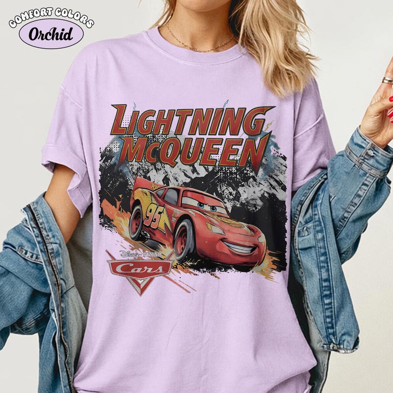 May include: A light purple short-sleeve t-shirt featuring a graphic of the red race car Lightning McQueen from the movie Cars. The text reads Lightning McQueen and includes the Cars logo. A Comfort Colors Orchid label is visible in the corner.