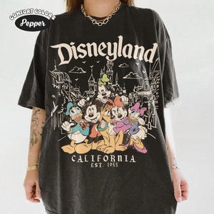 May include: Black t-shirt featuring the word "Disneyland" above a graphic of Disney characters, including Mickey Mouse, Donald Duck, and Goofy. The text "CALIFORNIA EST. 1955" is printed below the characters. The shirt has a vintage look.