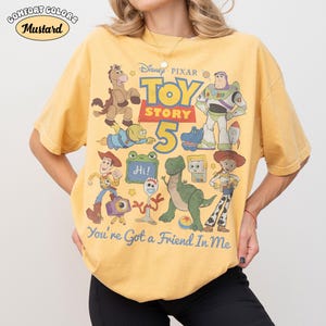 May include: Mustard-colored t-shirt featuring a graphic of Toy Story characters, including Woody, Buzz Lightyear, and Rex. The text "TOY STORY 5" and "You're Got a Friend In Me" are also displayed. The shirt has a Comfort Colors label.