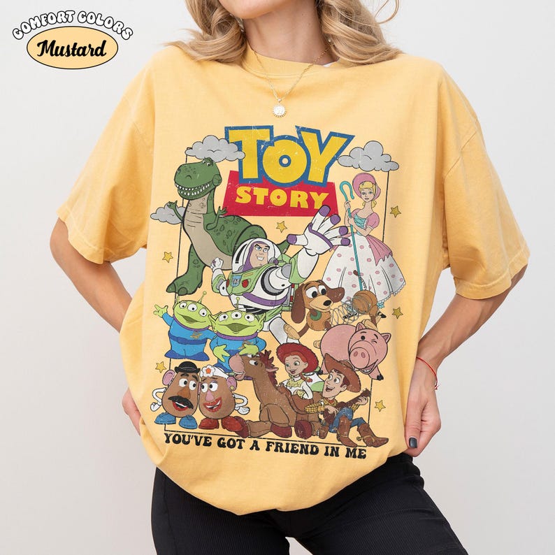May include: A mustard yellow short-sleeve t-shirt featuring a graphic print of Toy Story characters including Buzz Lightyear, Woody, Rex, and others. The text TOY STORY and YOU'VE GOT A FRIEND IN ME are printed in bold letters on the front of the shirt.