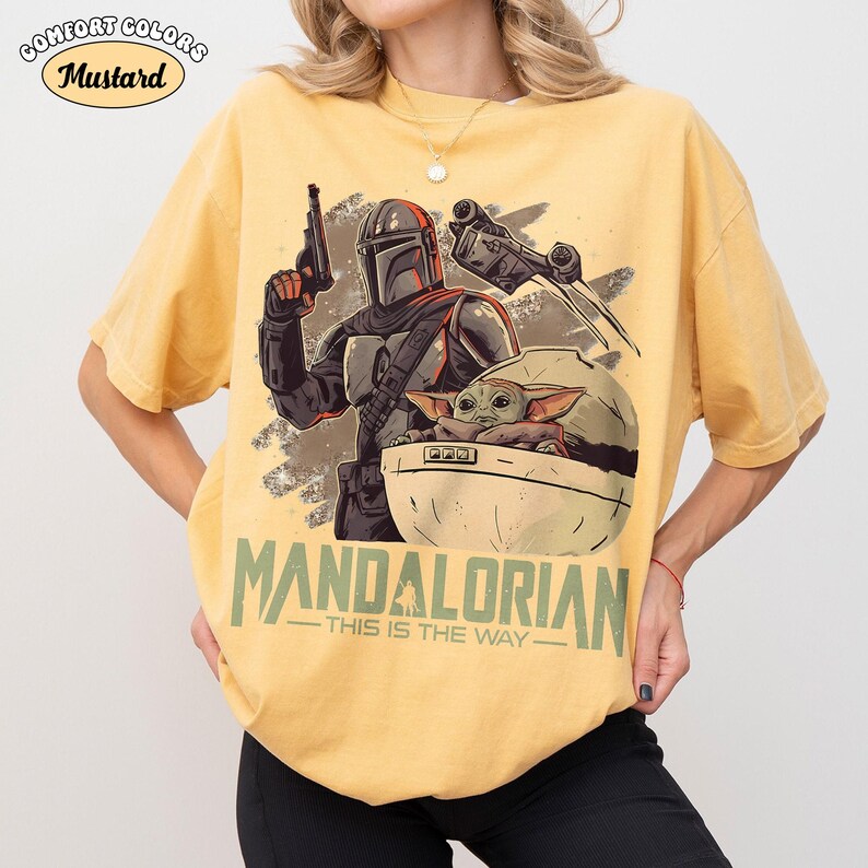 May include: A mustard yellow short-sleeve t-shirt featuring a graphic print of a Mandalorian character holding a blaster, a small creature in a floating pod, and a spacecraft. The text MANDALORIAN THIS IS THE WAY is printed in green below.