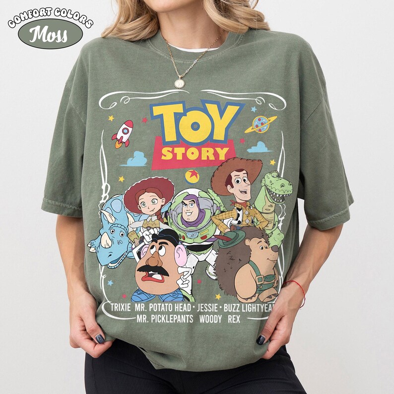 May include: A moss green Comfort Colors t-shirt featuring the Toy Story logo and characters. The design includes the words "Toy Story" in yellow, and the names of the characters: Trixie, Mr. Potato Head, Jessie, Buzz Lightyear, Mr. Picklepants, Woody Rex.