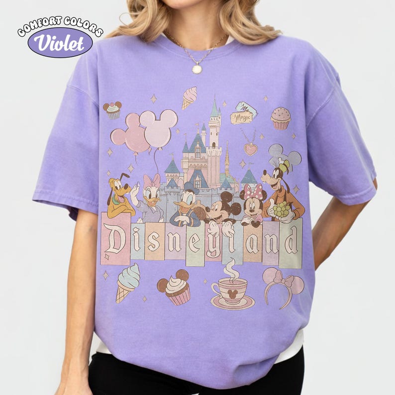 May include: A Comfort Colors violet t-shirt featuring a Disneyland graphic. The design includes cartoon characters like Mickey and Minnie Mouse, Donald and Daisy Duck, and Pluto, along with a castle, balloons, and the word "Disneyland".