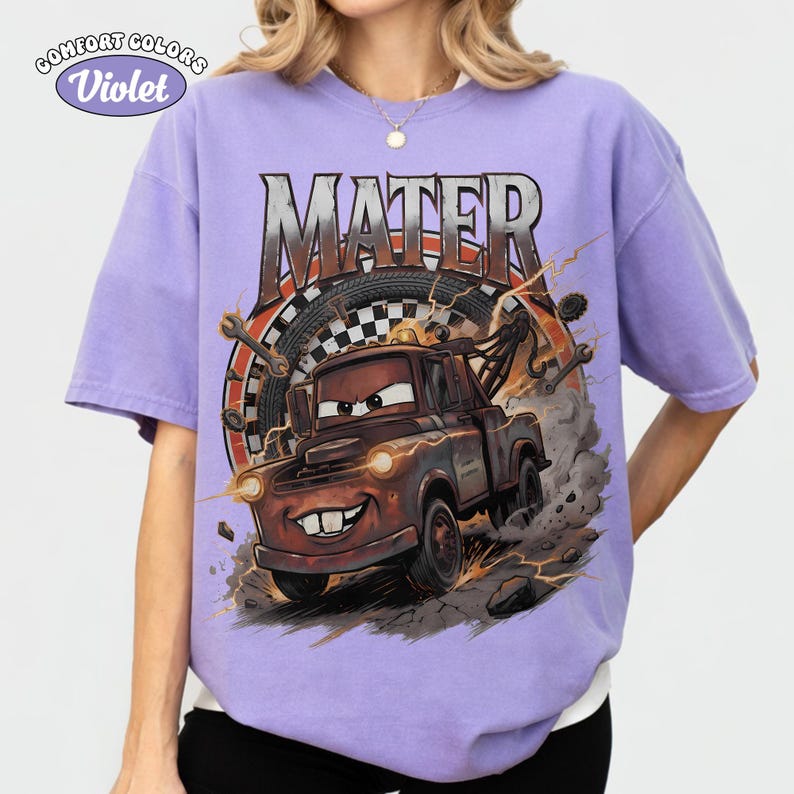 May include: Violet Comfort Colors t-shirt featuring a graphic of Mater from the movie Cars. The design includes the word "MATER" above the cartoon tow truck, with a checkered flag and tire background. The shirt is a light purple color.