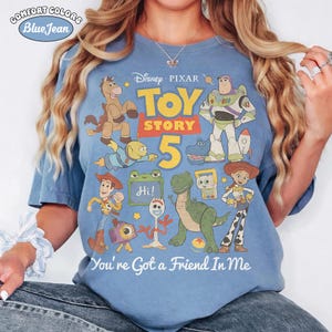 May include: Blue Comfort Colors t-shirt featuring a colorful graphic of characters from Disney Pixar's Toy Story 5. The design includes Woody, Buzz Lightyear, and other characters, with the text "You're Got a Friend In Me".