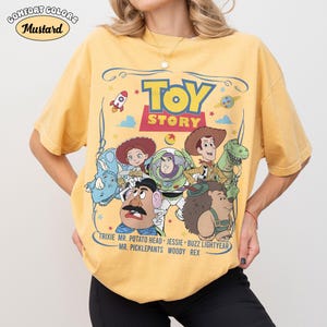 May include: Mustard-colored t-shirt featuring the Toy Story logo and characters. The graphic includes Buzz Lightyear, Woody, and other characters from the animated film. The shirt has the words "COMFORT COLORS" and "Mustard" printed on it.