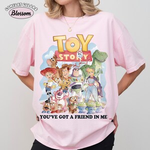 Puede incluir: Camiseta rosa claro Comfort Colors con un gr&aacute;fico colorido de personajes de Toy Story. El dise&ntilde;o incluye a Woody, Buzz Lightyear y otros personajes, con el texto "YOU'VE GOT A FRIEND IN ME".