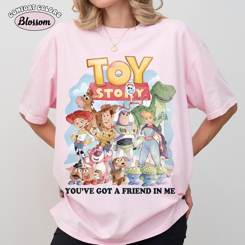 Puede incluir: Camiseta rosa claro Comfort Colors con un gr&aacute;fico colorido de personajes de Toy Story. El dise&ntilde;o incluye a Woody, Buzz Lightyear y otros personajes, con el texto "YOU'VE GOT A FRIEND IN ME".