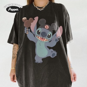 May include: A charcoal black short-sleeve t-shirt featuring a graphic of the character Stitch wearing a black cap with mouse ears and holding a brown ice cream treat. The text Comfort Colors Pepper is printed in the upper left corner of the shirt.