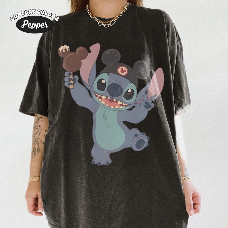 May include: A charcoal black short-sleeve t-shirt featuring a graphic of the character Stitch wearing a black cap with mouse ears and holding a brown ice cream treat. The text Comfort Colors Pepper is printed in the upper left corner of the shirt.