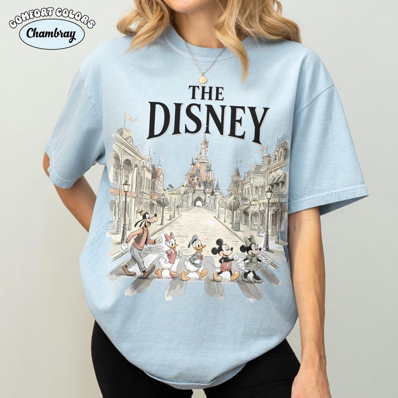 May include: Light blue t-shirt featuring the text "THE DISNEY" above a graphic of Disney characters crossing a street in front of a castle. The shirt has short sleeves and a crew neck.