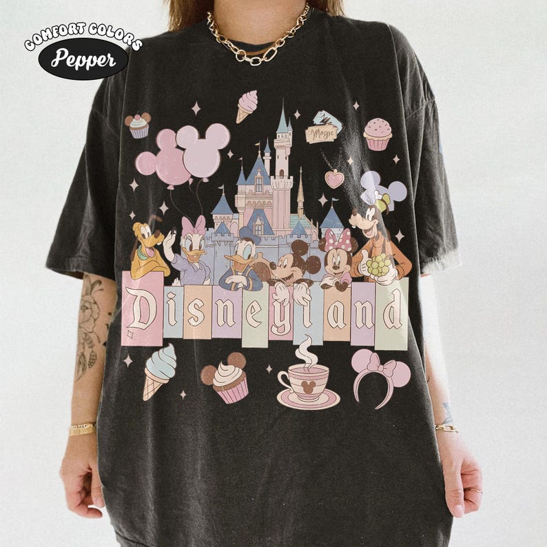 May include: Black t-shirt featuring a Disneyland graphic. The design includes the Disneyland castle, Mickey and Minnie Mouse, Donald and Daisy Duck, Goofy, and Pluto. The shirt also has pink balloons, ice cream cones, cupcakes, and the word "Disneyland".
