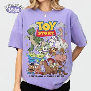 May include: A violet short-sleeve t-shirt featuring a graphic print of Toy Story characters including Buzz Lightyear, Woody, Rex, and Bo Peep. The text reads TOY STORY and YOU'VE GOT A FRIEND IN ME. The shirt has a Comfort Colors Violet label.