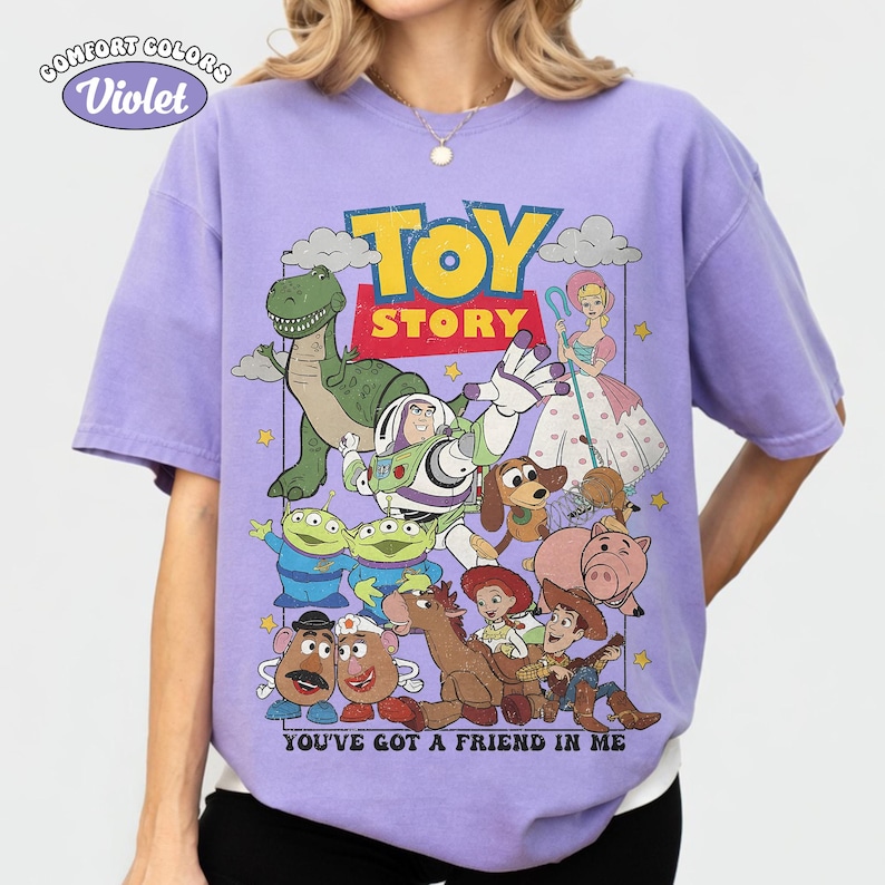 May include: A violet short-sleeve t-shirt featuring a graphic print of Toy Story characters including Buzz Lightyear, Woody, Rex, and Bo Peep. The text reads TOY STORY and YOU'VE GOT A FRIEND IN ME. The shirt has a Comfort Colors Violet label.