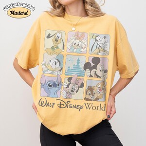 Op de afbeelding: Een mosterdgeel T-shirt met korte mouwen voorzien van een raster van negen vierkanten met diverse Disney-figuren, waaronder Mickey Mouse, Minnie Mouse, Donald Duck, Daisy Duck, Goofy, Pluto, Stitch, Knabbel en Babbel, en de tekst Walt Disney World.