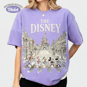 May include: Violet Comfort Colors t-shirt featuring a Disney design. The design includes the text "THE DISNEY" above an illustration of Disney characters crossing a street in front of a castle.