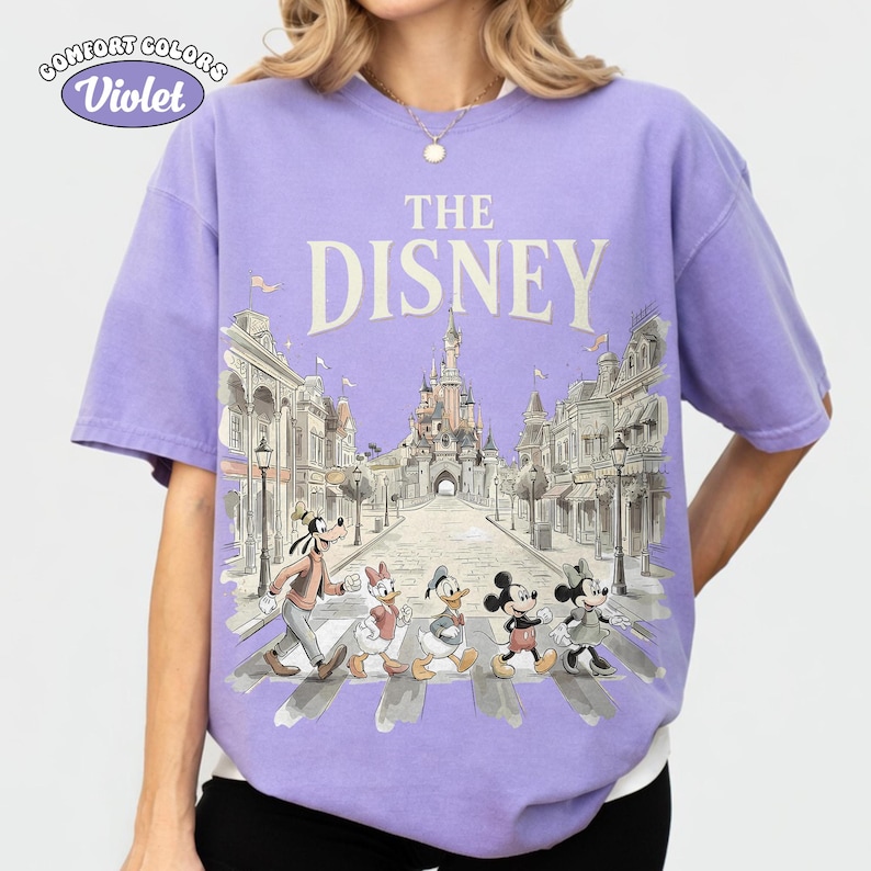 May include: Violet Comfort Colors t-shirt featuring a Disney design. The design includes the text "THE DISNEY" above an illustration of Disney characters crossing a street in front of a castle.