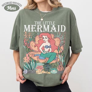 May include: Olive green t-shirt featuring a graphic of Ariel from The Little Mermaid. The design includes the text "THE LITTLE MERMAID" above an illustration of Ariel in a seashell, surrounded by coral and seaweed. The shirt has the Comfort Colors logo.