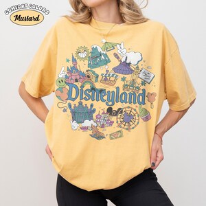 May include: Mustard yellow t-shirt featuring a Disneyland graphic. The design includes park icons such as a castle, a Ferris wheel, and the Disneyland logo. The shirt has short sleeves and a round neck.