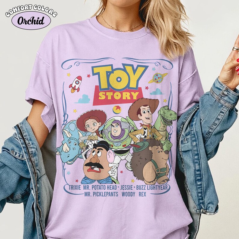 May include: A lilac Comfort Colors t-shirt featuring the Toy Story movie characters. The graphic includes the words "TOY STORY" in yellow, and the characters Woody, Buzz Lightyear, and Mr. Potato Head. The shirt also lists character names.